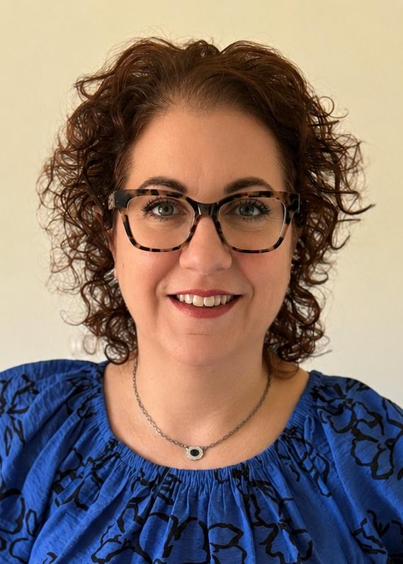 A woman with curly hair, a blue patterned shirt and glasses smiles at the camera.